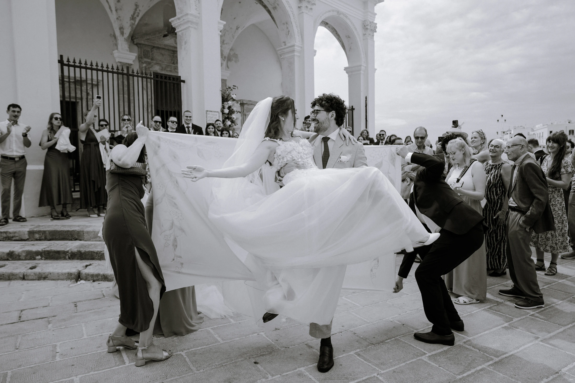 Lo sposo solleva la sposa all'uscita dalla chiesa tra gli invitati, in bianco e nero